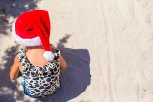 menina bonitinha com chapéu de Papai Noel vermelho na praia branca ao ar livre foto