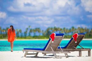 duas espreguiçadeiras com chapéus de papai noel vermelhos na praia branca tropical com água azul-turquesa foto