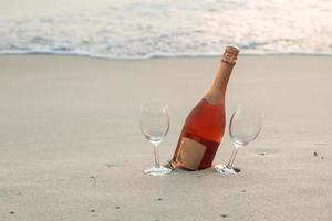 garrafa de vinho tinto e dois copos na praia de areia branca foto