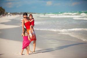 jovem mãe e duas filhas de moda caminhando na praia exótica em dia ensolarado foto