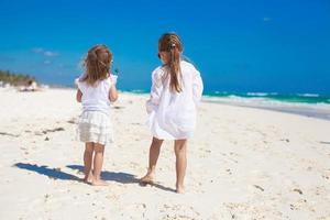 vista traseira de duas irmãzinhas em roupas brancas se divertindo na praia de areia tropical foto
