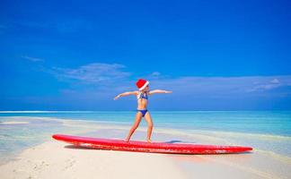 adorável garotinha com chapéu de Papai Noel pratica posição de surf na prancha de surf durante as férias foto