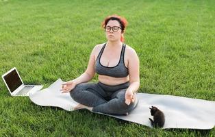 mulher de positividade corporal plus size fazendo meditação no tapete de ioga com gatinho no quintal - conceito de bem-estar e saúde mental foto