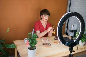 blogueira de beleza feminina de meia-idade ou influenciadora com vídeo de gravação de produto de maquiagem no quarto em casa. usando lâmpada de anel e smartphone foto