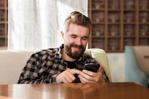 homem sorridente usando smartphone na cafeteria moderna, ele conversando mensagens on-line no celular. redes sociais e conceito da geração do milênio foto