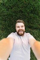 jovem com barba e estilo de cabelo da moda deitado na grama tomando selfie - segurando o smartphone ou tablet e olhando para a câmera. foto