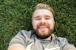 jovem com barba e estilo de cabelo da moda deitado na grama tomando selfie - segurando o smartphone ou tablet e olhando para a câmera. foto