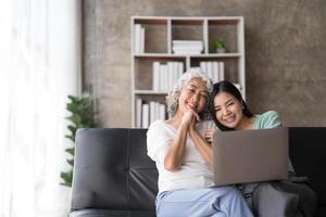 mãe madura, filha crescida, sente-se no sofá na sala de estar, segure no colo do pc, olhando para a tela, assistindo a um filme, usando o dispositivo, faça uma chamada de vídeo, fazendo compras via site de comércio eletrônico, a menina ensina o conceito de mãe foto