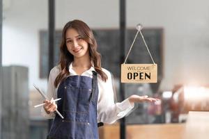 bela jovem asiática barista no avental segurando o tablet e em frente à porta do café com placa aberta. conceito de inicialização do proprietário do negócio. foto