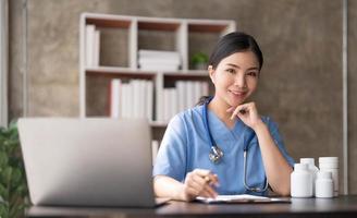 jovem médica asiática bonita sorrindo usando o trabalho com um computador portátil e ela escrevendo algo na papelada ou na prancheta papel branco no escritório da mesa do hospital, conceito médico de saúde foto