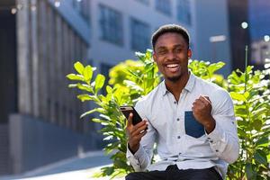 homem afro-americano feliz em roupas casuais, sentado no banco ao ar livre com telefone móvel foto