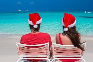 casal jovem feliz em chapéus de papai noel vermelhos sentados em cadeiras de praia foto