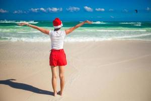 vista traseira da jovem com chapéu de Papai Noel andando espalhou as mãos na praia branca foto