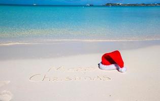 feliz natal escrito na areia branca da praia tropical com chapéu de natal foto