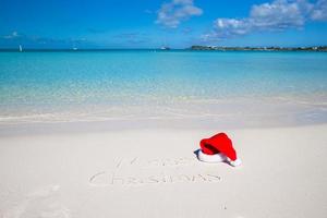 feliz natal escrito na areia branca da praia tropical com chapéu de natal foto