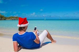 vista traseira do jovem com chapéu de Papai Noel com telefone na praia tropical do Caribe foto