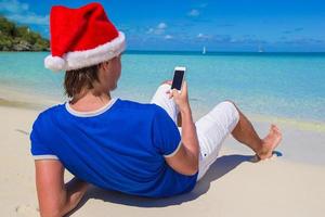 vista traseira do jovem com chapéu de Papai Noel com telefone na praia tropical do Caribe foto