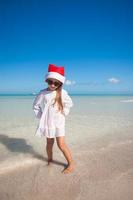 menina bonitinha de chapéu vermelho de papai noel e óculos de sol na praia foto