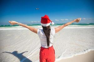 vista traseira da linda garota com chapéu de Papai Noel andando como um pássaro na praia branca foto