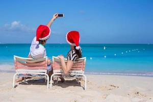 jovem casal feliz em chapéus de papai noel vermelhos tirando uma foto no celular