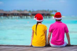 vista traseira do jovem casal em chapéus de papai noel no cais de madeira nas férias de natal foto