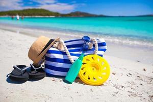 bolsa listrada, chapéu de palha, protetor solar e toalha na praia tropical branca foto