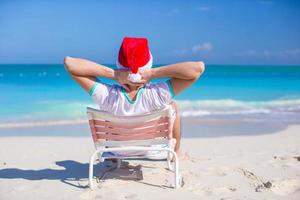 vista traseira do jovem com chapéu de Papai Noel relaxar na cadeira de praia foto