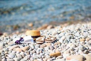 chapéu de praia no livro aberto com protetor solar e sapatos na praia de seixos foto