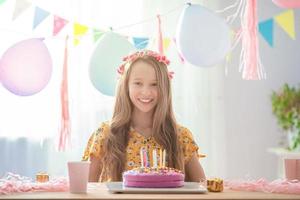 garota caucasiana está sonhando sorrindo e olhando para o bolo de arco-íris de aniversário. fundo colorido festivo com balões. festa de aniversário e conceito de desejos. foto