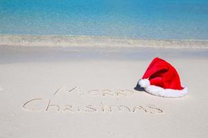 feliz natal escrito na areia branca da praia tropical com chapéu de natal foto