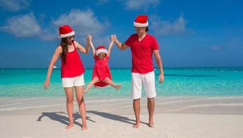 família feliz de três em chapéus de natal se divertindo na praia branca foto