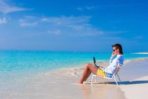 jovem com laptop e celular na praia tropical foto