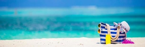 bolsa listrada, toalha azul, óculos de sol, garrafa de protetor solar e maiô na praia branca foto