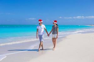 jovem casal feliz em chapéus de papai noel vermelhos andando na praia de areia tropical foto