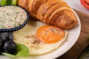 croissant, ovo frito, molho para salada, uvas pretas e tomates foto