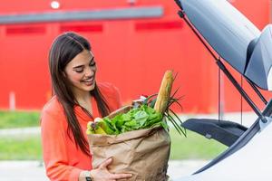 morena caucasiana vai segurando sacos de papel com produtos alimentícios. jovem mulher colocando pacote com mantimentos e legumes na mala do carro. mulher caucasiana atraente fazendo compras no shopping ou mercearia foto