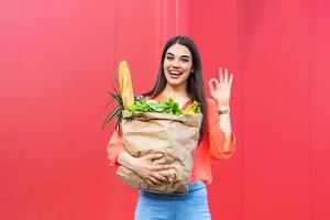 jovem mulher bonita segurando o saco de papel de mantimentos do supermercado com um sorriso no rosto mostrando ok sinal de pé e sorrindo com um sorriso confiante foto