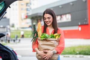 mulher colocando suas sacolas de compras no carro no estacionamento do shopping. ela fez algumas compras leves, principalmente comida. jovem mulher bonita feliz muda a compra do carrinho de compras no porta-malas foto