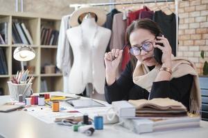 designer de moda feminina asiática de meia-idade trabalha em estúdio falando no celular sobre ideias e desenhando esboços para pedidos de coleção de design de vestidos. boutique profissional sob medida empresário. foto