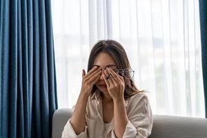 jovem esfregando os olhos sente dor e tira os óculos sentada no sofá e descansa os olhos em casa, problema de visão foto