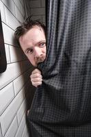 homem molhado e alarmado olha para fora do chuveiro, morde a cortina com entusiasmo e desvia o olhar consternado. foto