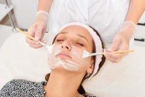 cosmetologista aplicando máscara de creme cosmético no rosto de mulher para rejuvenescimento, procedimento em salão de beleza foto