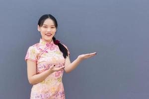 linda mulher asiática com cabelo comprido que usa vestido cheongsam rosa no tema do ano novo chinês enquanto sua mão mostra para apresentar algo e olha para a câmera sorrindo alegremente com o fundo cinza. foto