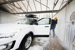 homem lavando carro suv americano de água de alta pressão com rack de teto na lavagem de autoatendimento em clima frio. foto