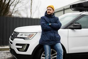 motorista de homem usa jaqueta e chapéu amarelo contra seu carro suv americano com rack de teto em tempo frio. foto