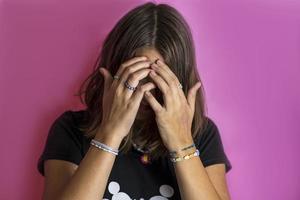 uma jovem adolescente cobre o rosto com as mãos, em um fundo rosa, a garota está chateada, confusa foto