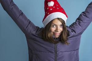 uma linda garota em um st. chapéu de nicholas e uma jaqueta quente sobre um fundo azul levanta as mãos, ano novo, natal foto