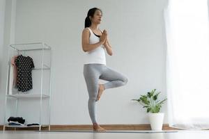 linda mulher asiática fazendo ioga em casa. as mulheres mantêm o equilíbrio em uma pose de árvore e praticam inspirando e expirando lentamente. conceito de saúde e fitness foto