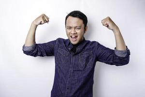 homem asiático animado, vestindo uma camisa azul, mostrando um gesto forte, levantando os braços e os músculos, sorrindo com orgulho foto