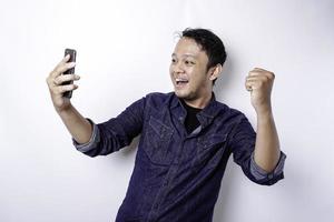 um jovem asiático com uma expressão feliz e bem-sucedida vestindo camisa azul e segurando seu telefone, isolado por fundo branco foto